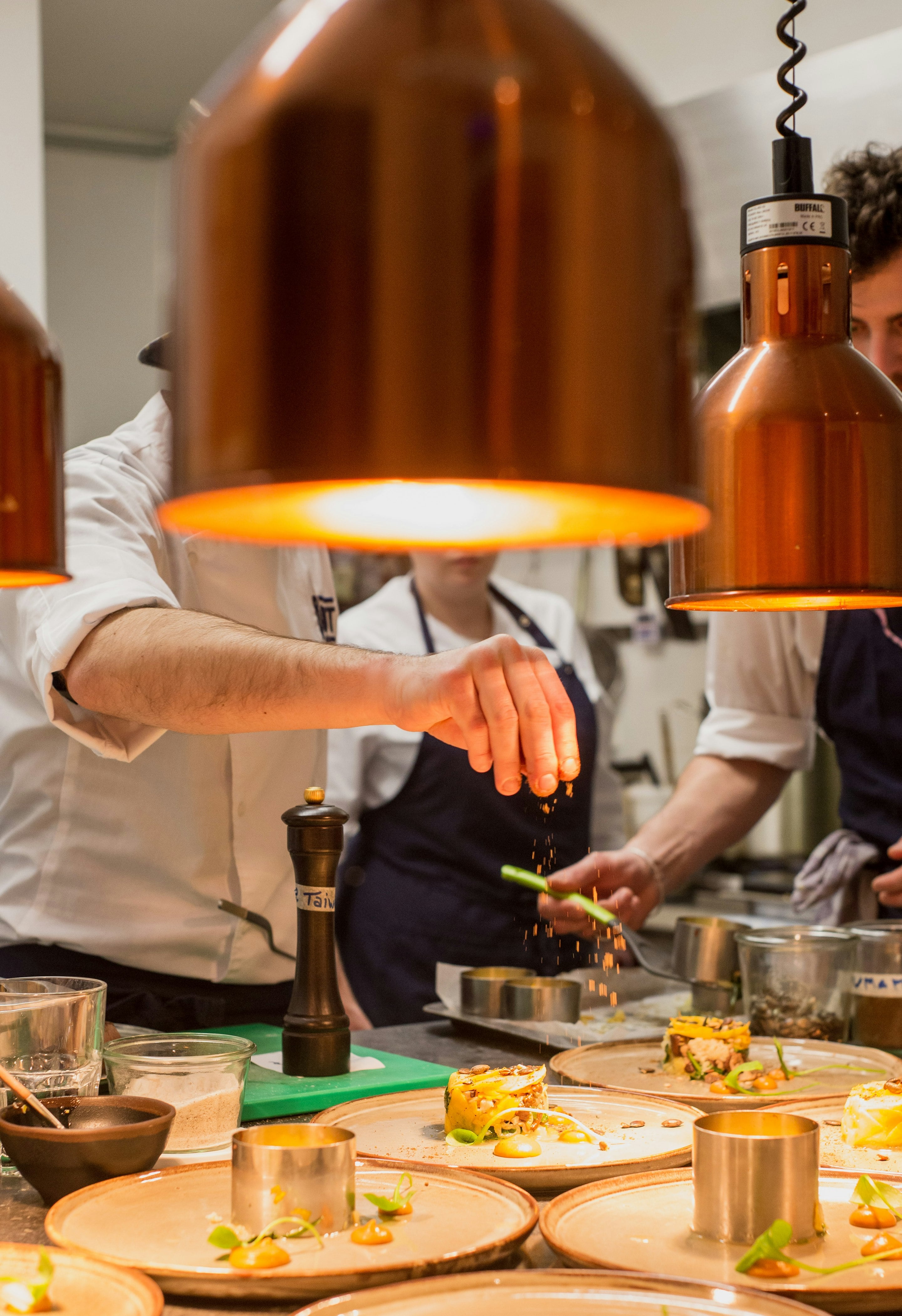 A chef seasons a row of plated dishes under copper heat lamps at the pass.