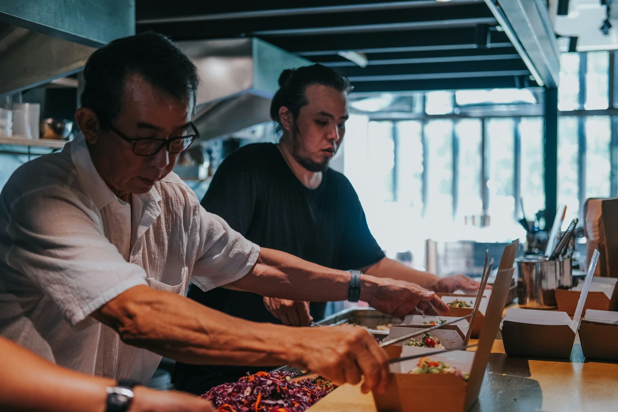 Two cooks packing orders side by side at a long kitchen counter.